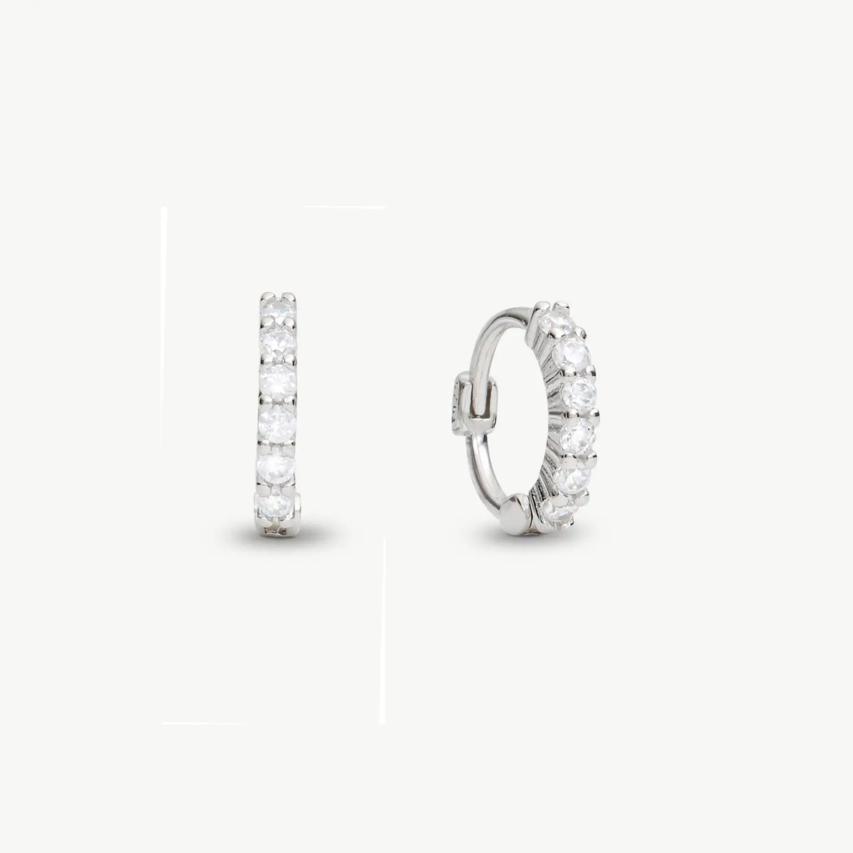 A pair of silver huggie hoop earrings with a row of clear round-cut stones, shown from front and side angles on a light gray background.