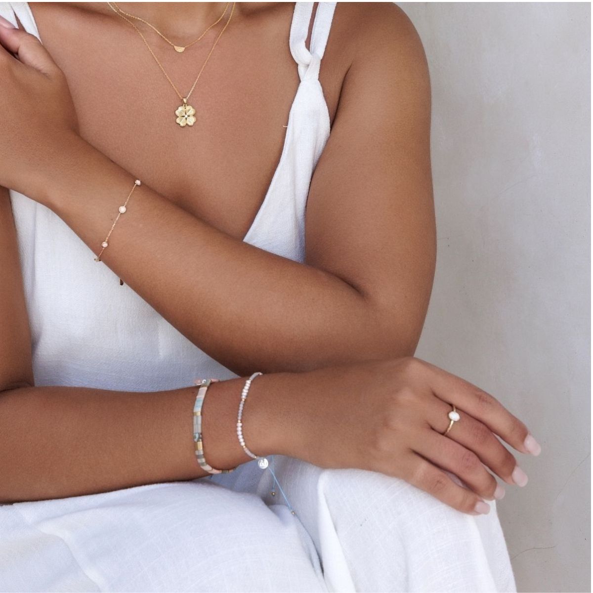 Close-up of a person wearing gold jewelry including a necklace, bracelet, and ring on a neutral background.