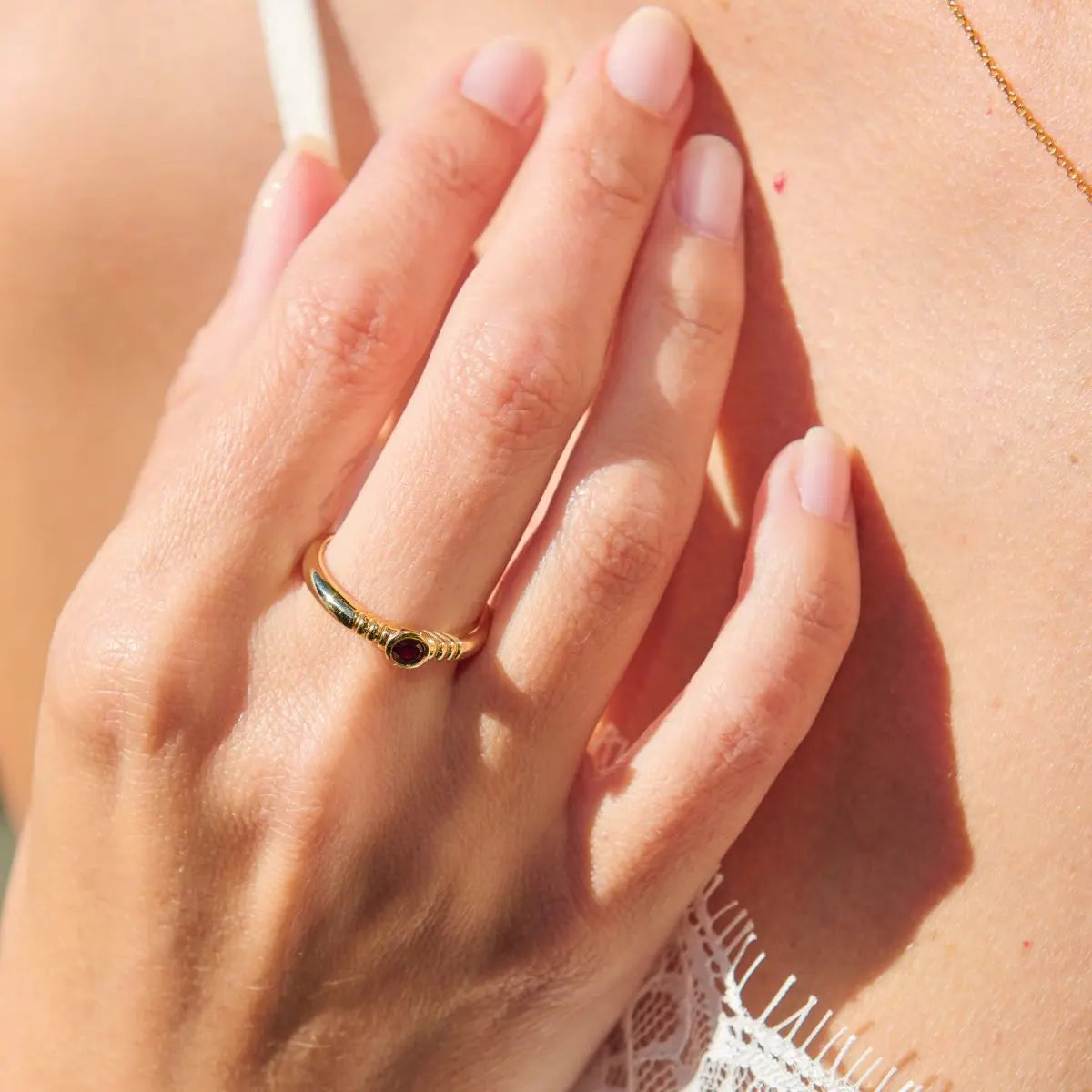 Close-up of a hand wearing a gold ring with a red gemstone, touching skin.