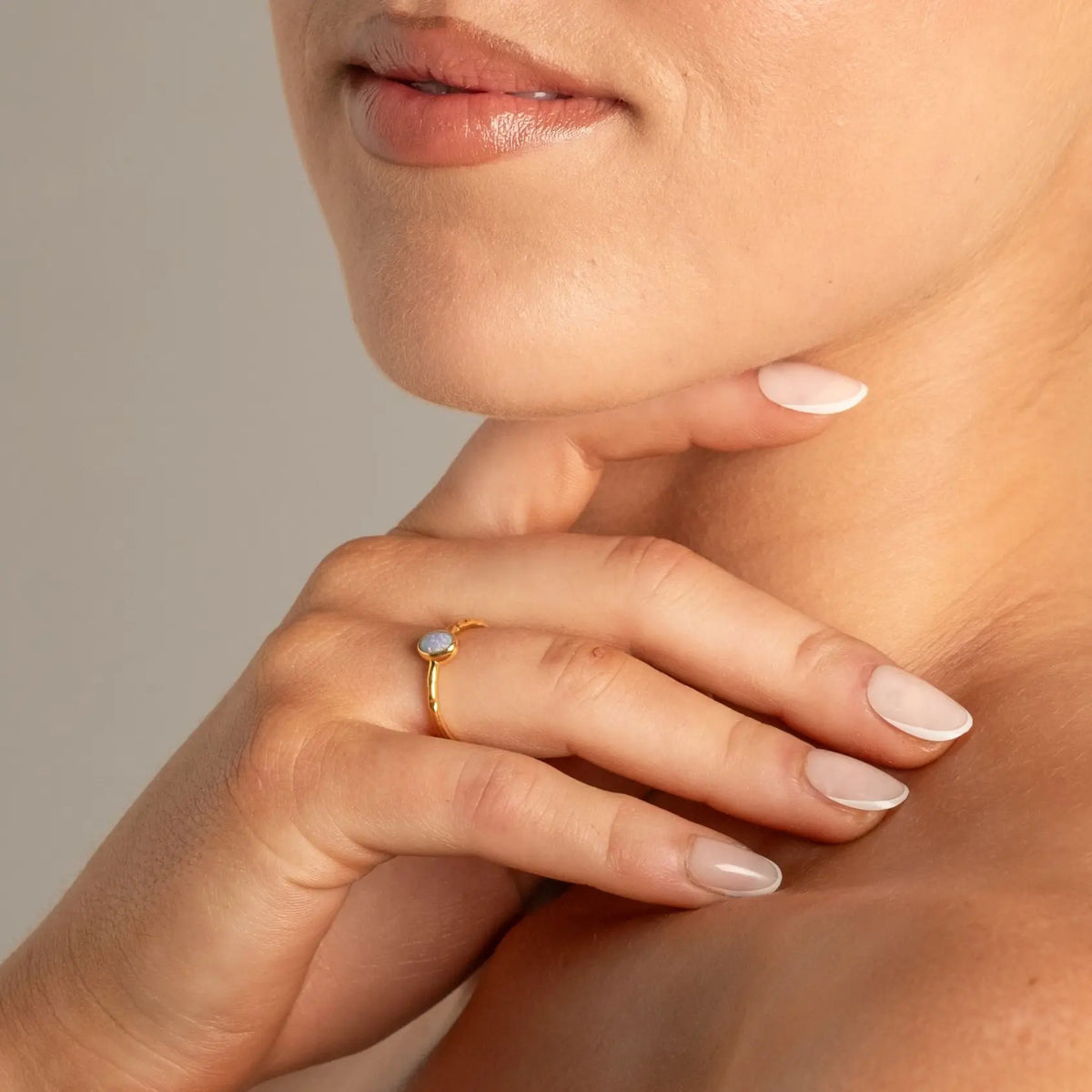 A close-up lifestyle shot of a woman's hand resting on her neck, wearing a delicate gold ring with a small oval light-colored gemstone.