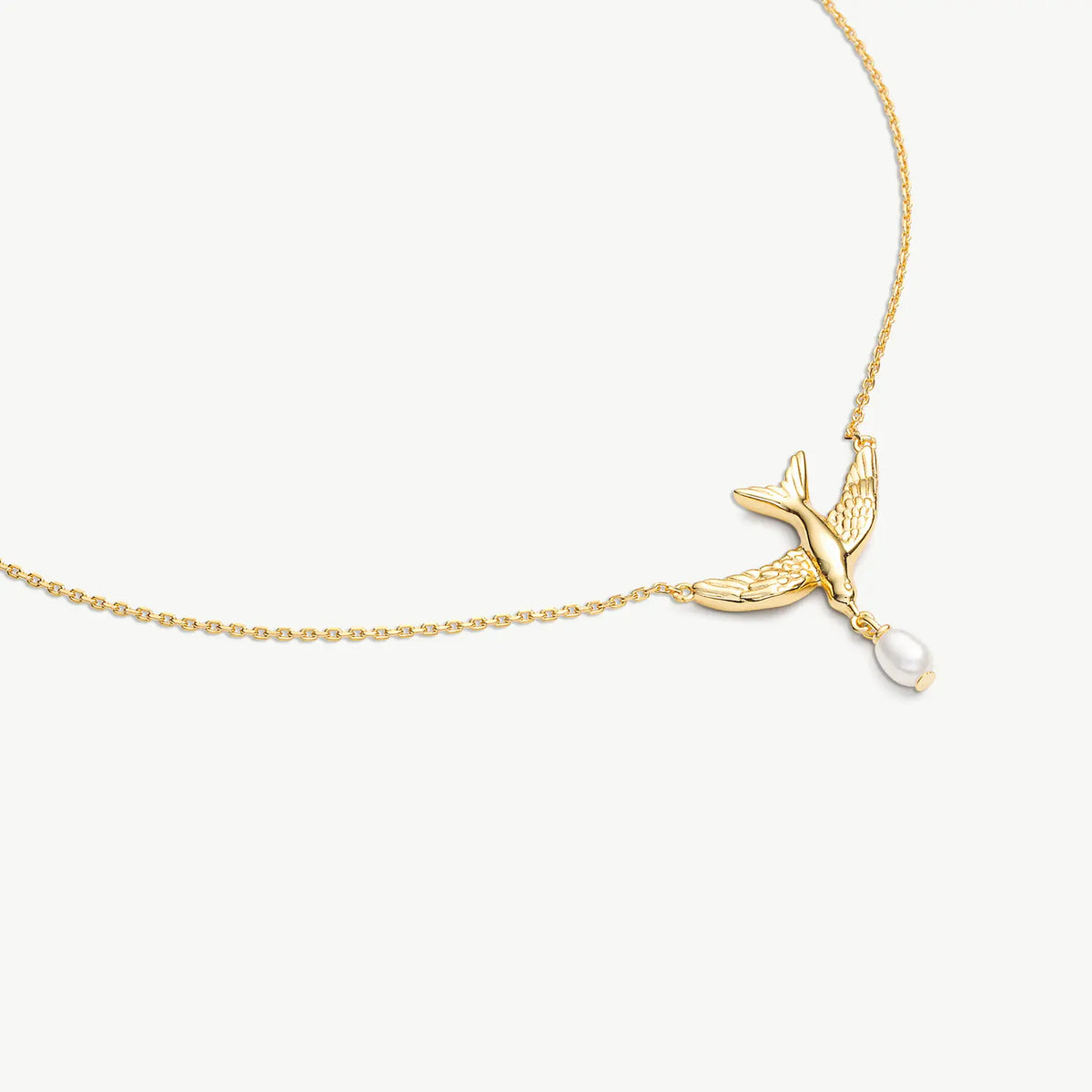 A delicate gold chain necklace featuring a small bird charm with a dangling white pearl, shown against a clean off-white background.