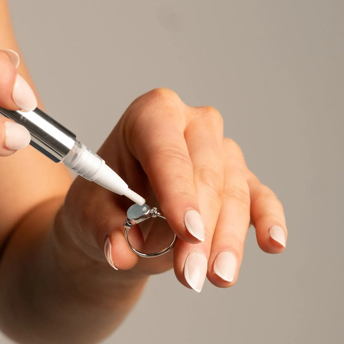 A close-up of a hand holding a silver ring while another hand applies a white liquid from a small applicator pen to the ring.