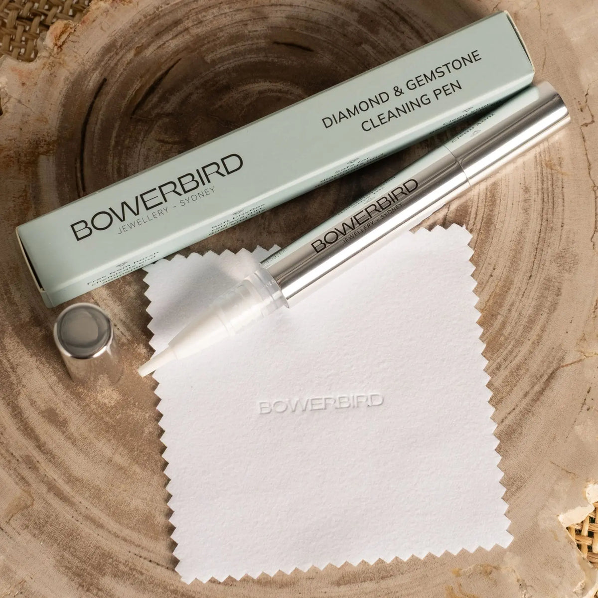 Bowerbird Diamond & Gemstone Cleaning Pen shown with its matching box, a white cleaning cloth, and cap on a rustic wood slice background.