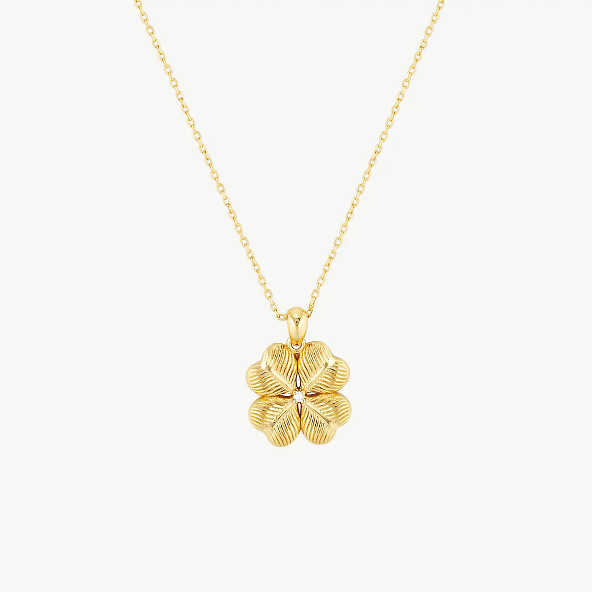 A ridged four-leaf clover charm with a cubic zirconia centred gemstone hanging from a fine yellow gold cable chain on a cream background