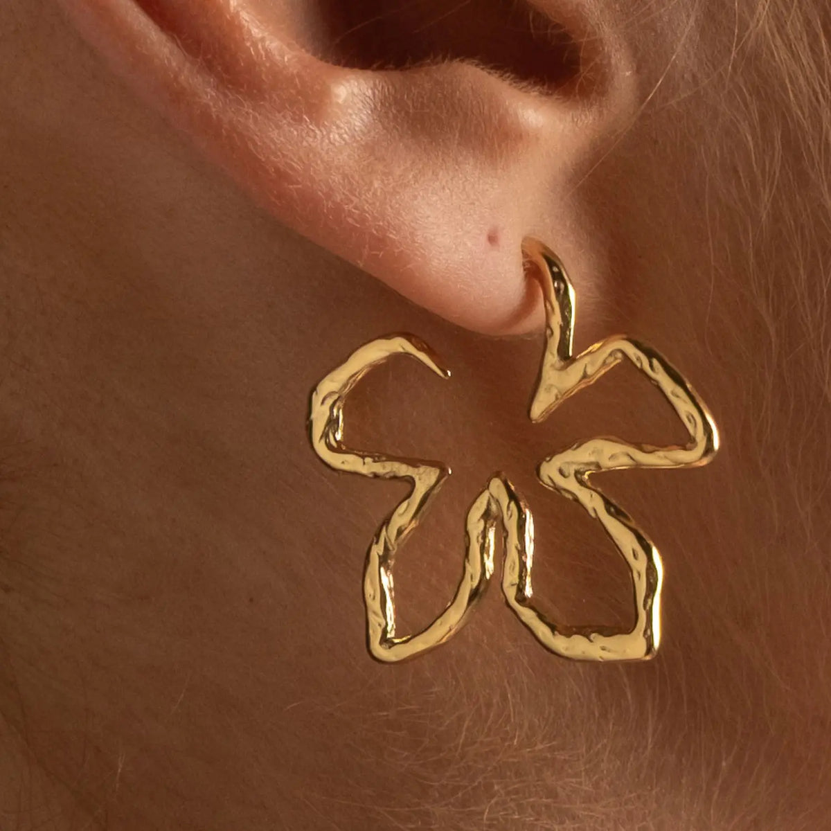 A close-up of a person's ear wearing a single textured gold hoop earring shaped like a stylized flower or abstract loop.