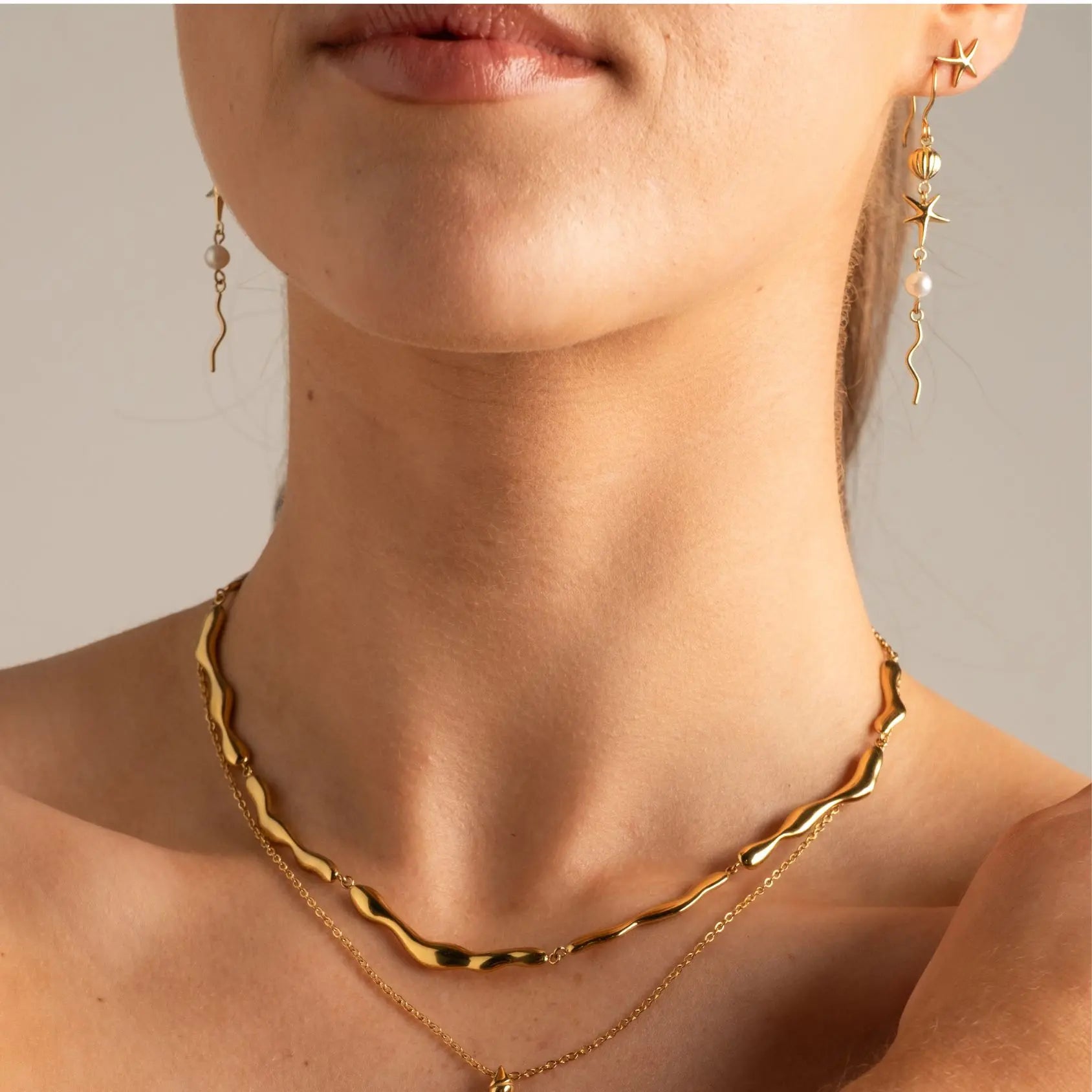 Close-up of a model wearing a gold wavy chain necklace layered with a delicate gold chain necklace, along with matching dangling earrings.