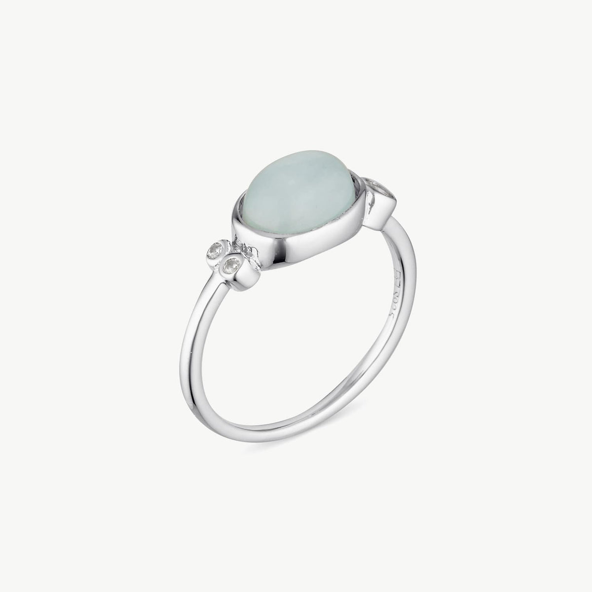 A silver ring featuring a smooth, oval-shaped pale blue stone and a small cluster of clear accent stones on the band, set against a plain off-white background.