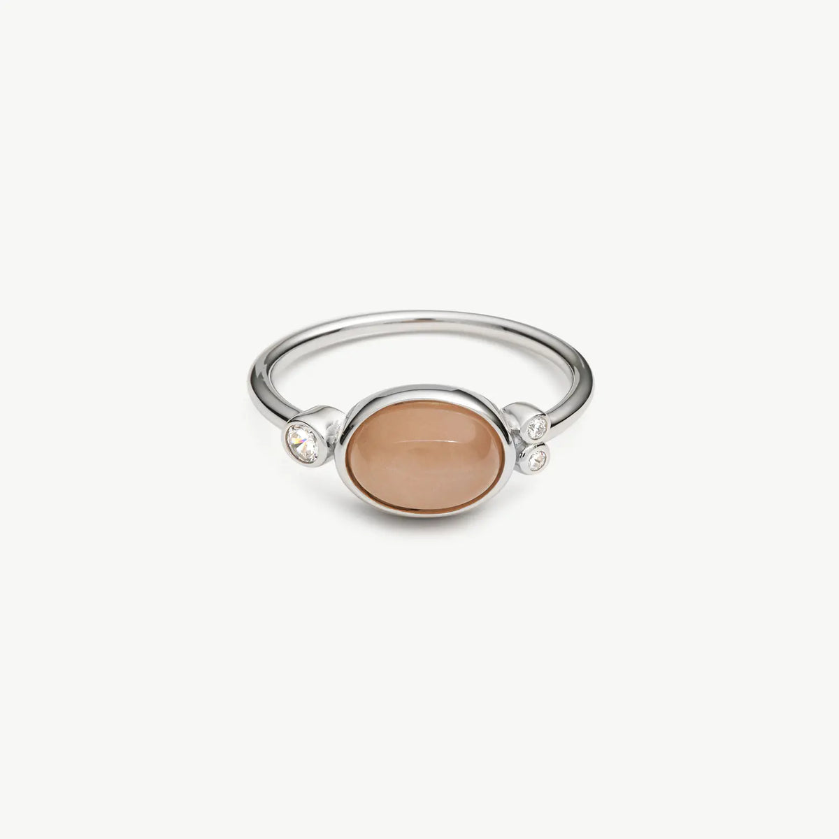 A silver ring featuring a smooth, oval-shaped peach-colored cabochon stone in the center, accented by two small clear crystals on either side of the band, set against a plain off-white background.