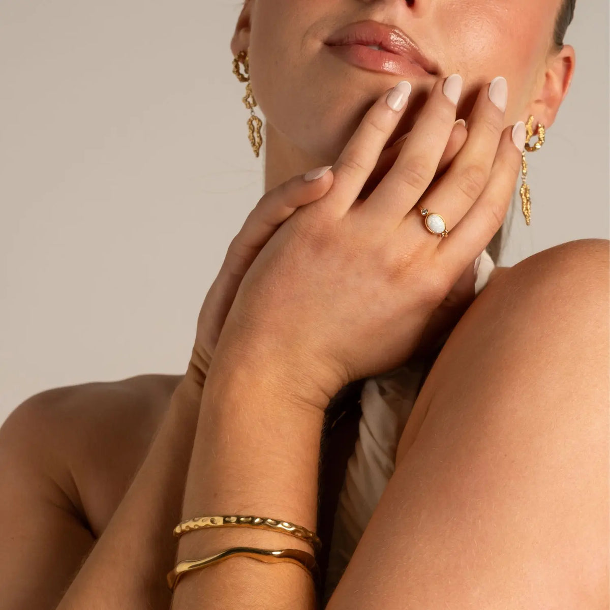 A close-up crop shot of a woman holding clasped hands resting against her chin. She's wearing long dangling organic drop gold earrings, a white cabochon gemstone ring on her finger, and two gold bangles.