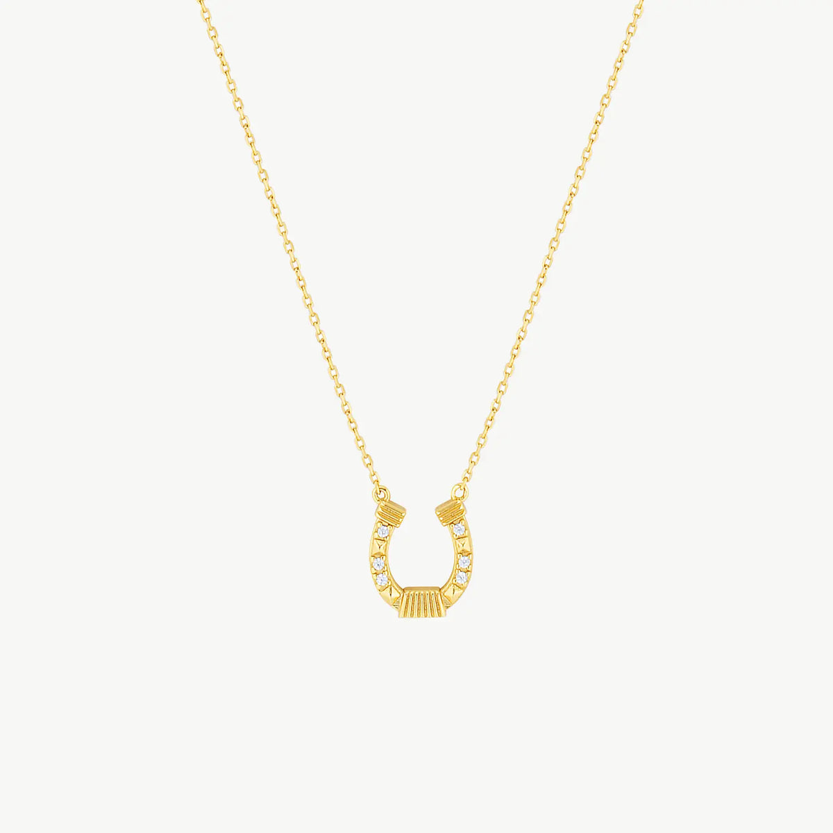 A yellow gold hanging horseshoe charm suspended from a fine gold cable chain with sparkling cubic zirconias on a cream background