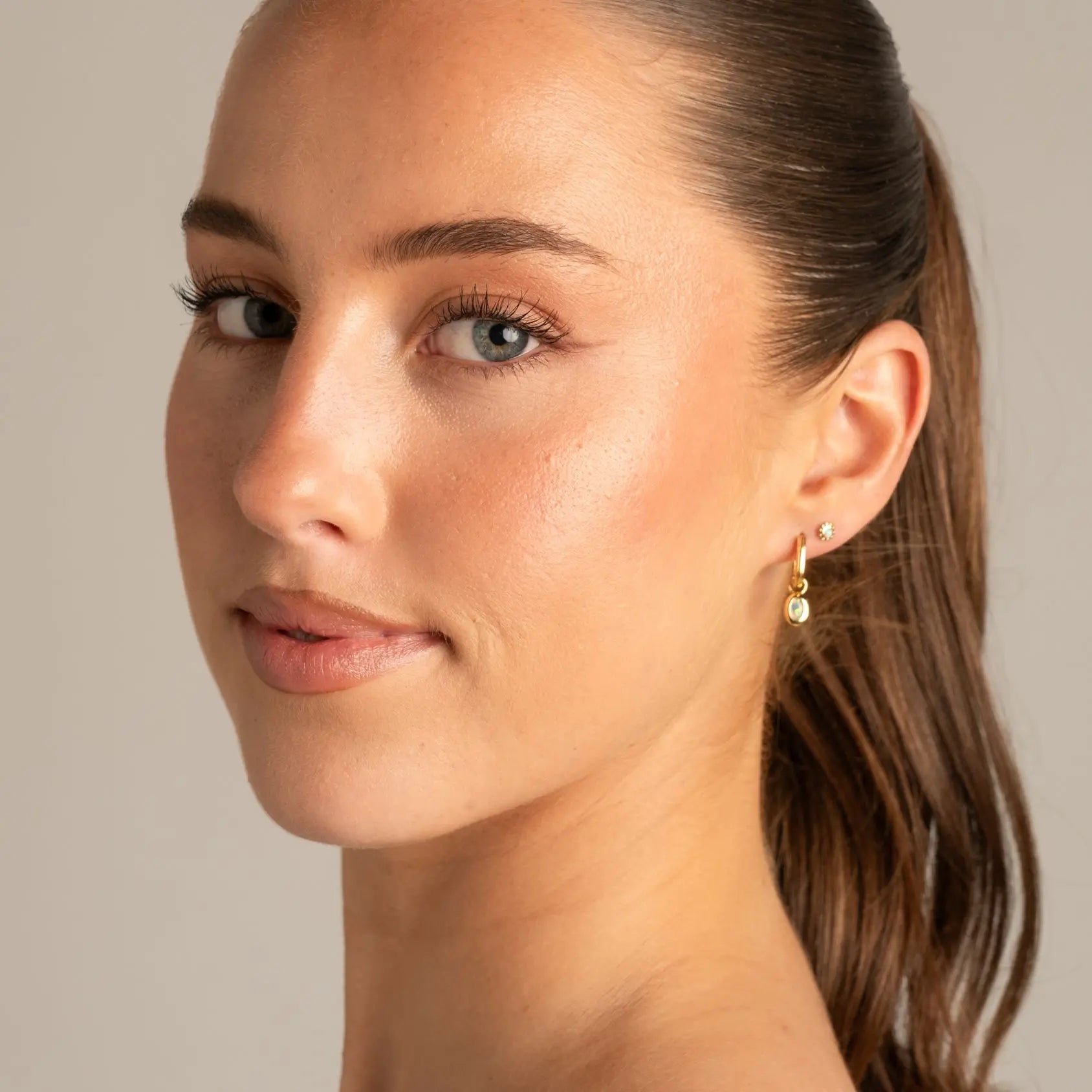 A close-up portrait of a woman with her hair in a ponytail, wearing a delicate gold dangle earring with a small clear stone, set against a soft neutral beige background.