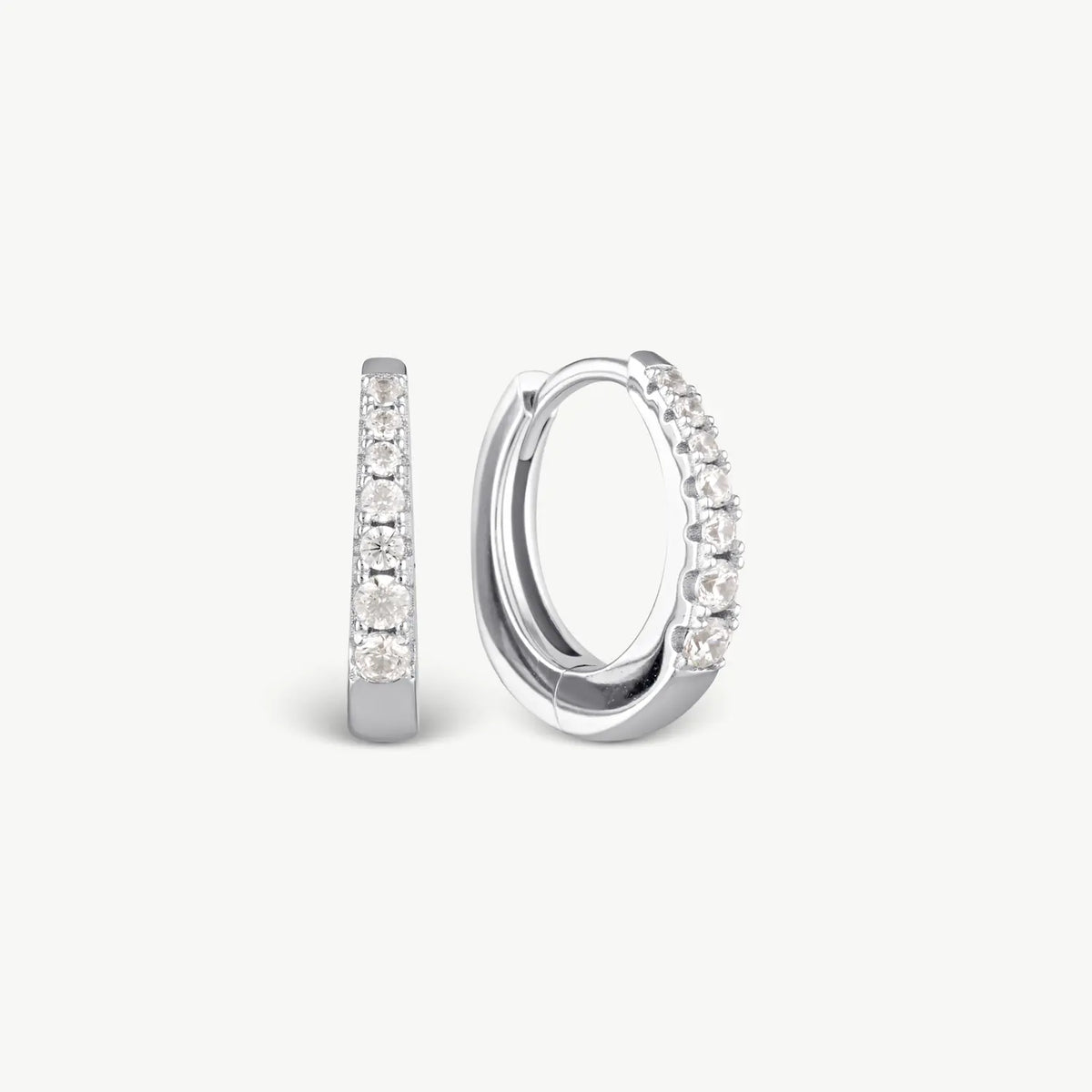 A pair of silver huggie hoop earrings with clear pavé-set stones, shown from side and front angles on a light gray background.