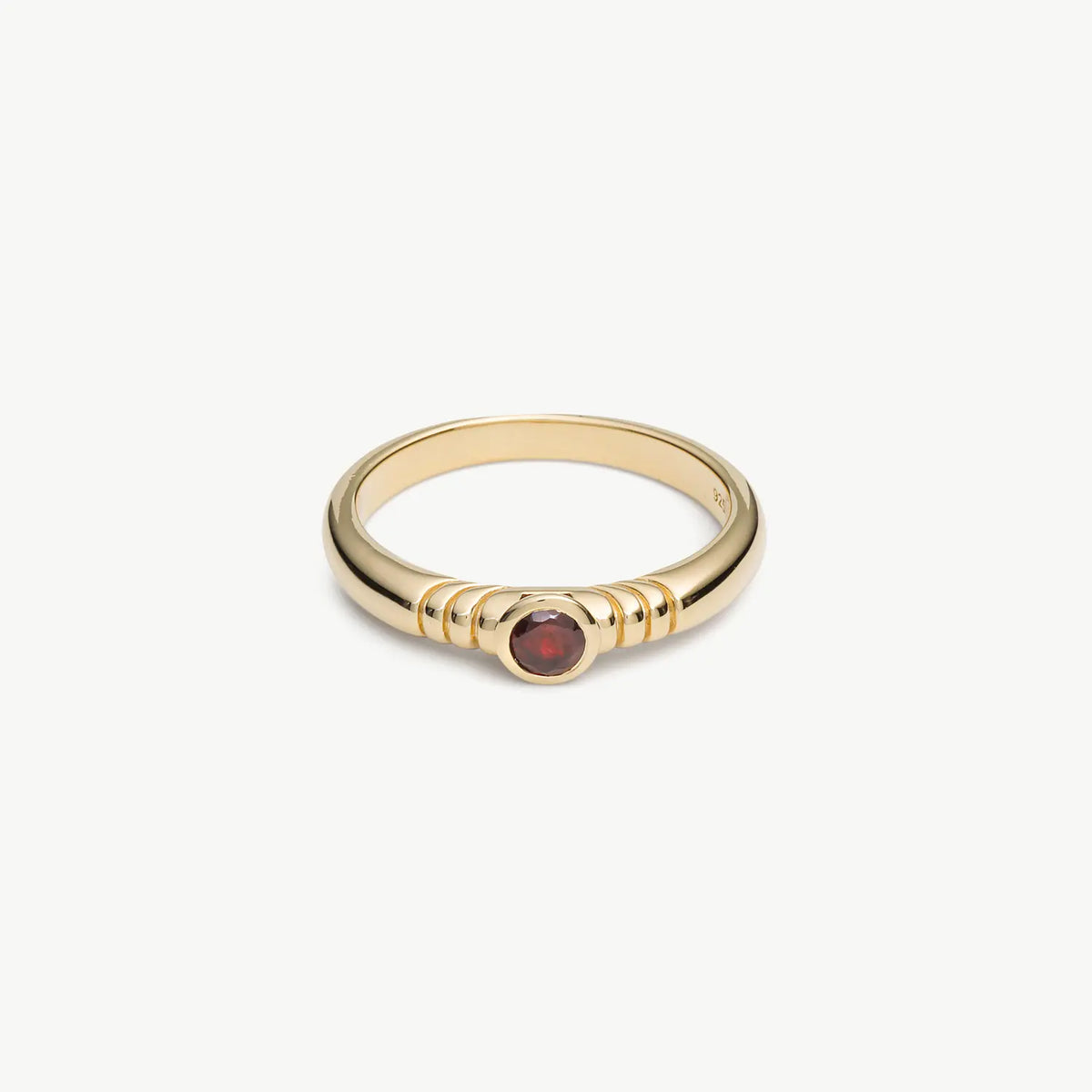 A gold ring with a textured band and a small round red gemstone in the center, shown on a clean off-white background.