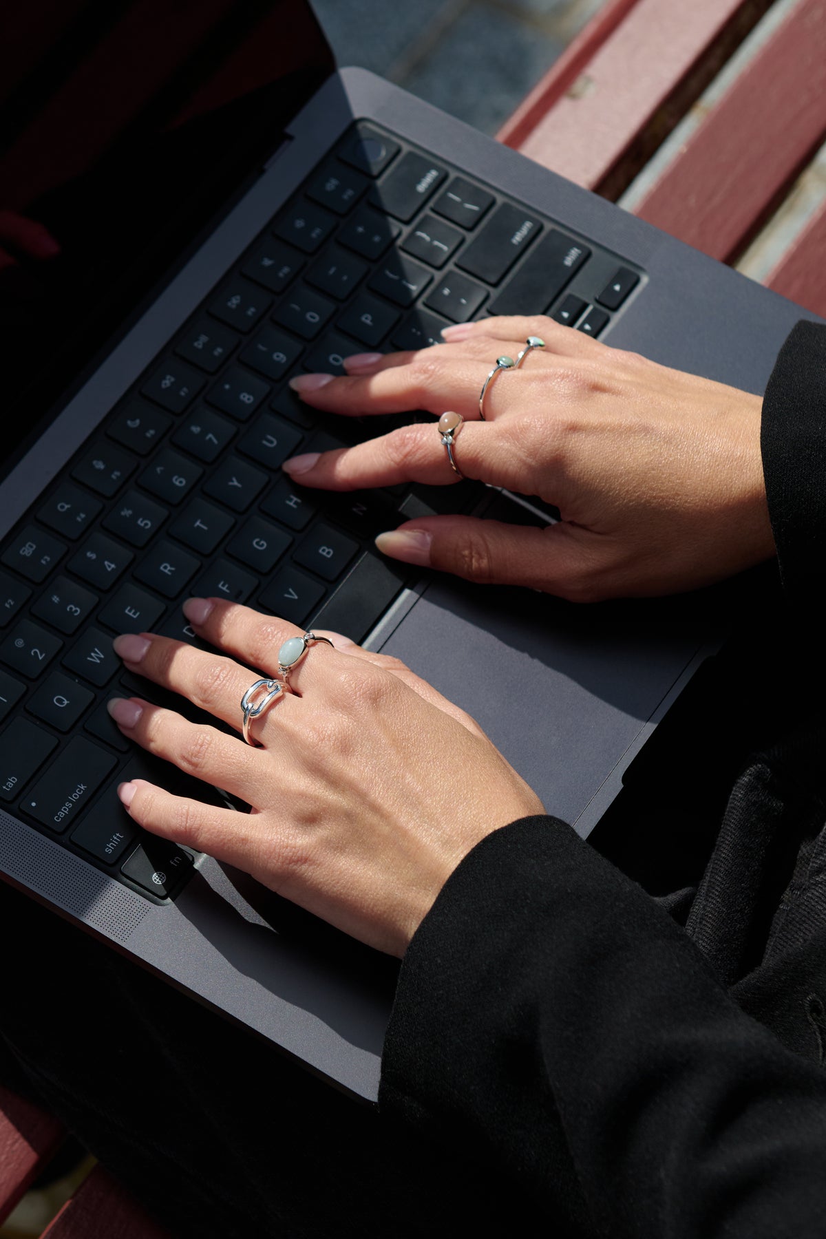 A close-up lifestyle shot of a person's hands typing on a laptop keyboard, wearing multiple silver rings with light blue and pink stones.