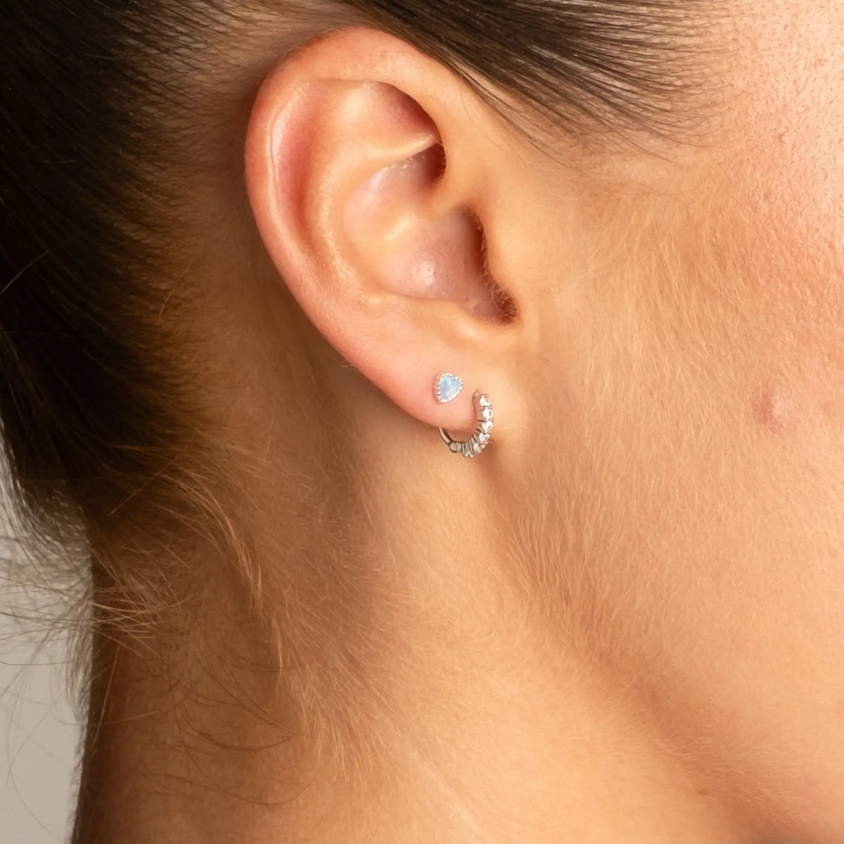 A close-up of a person's ear wearing a small silver crescent huggie hoop earring adorned with clear stones and a matching round stud earring.