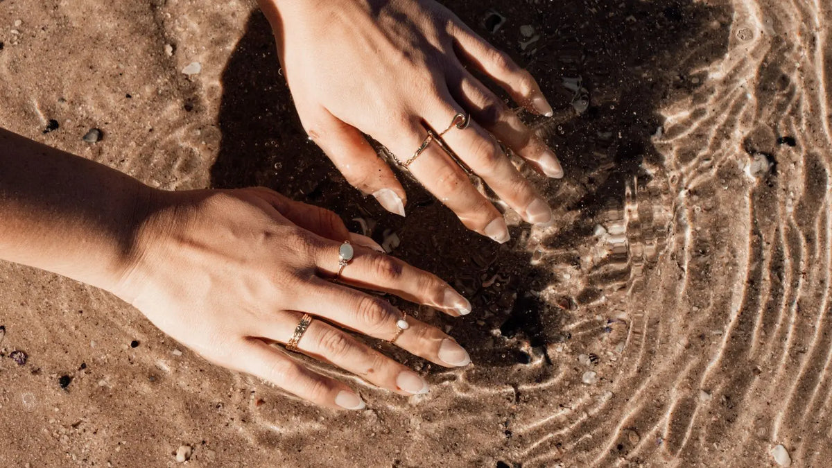 Two hands dipping into water at the beach with a range of different rings on fingers 