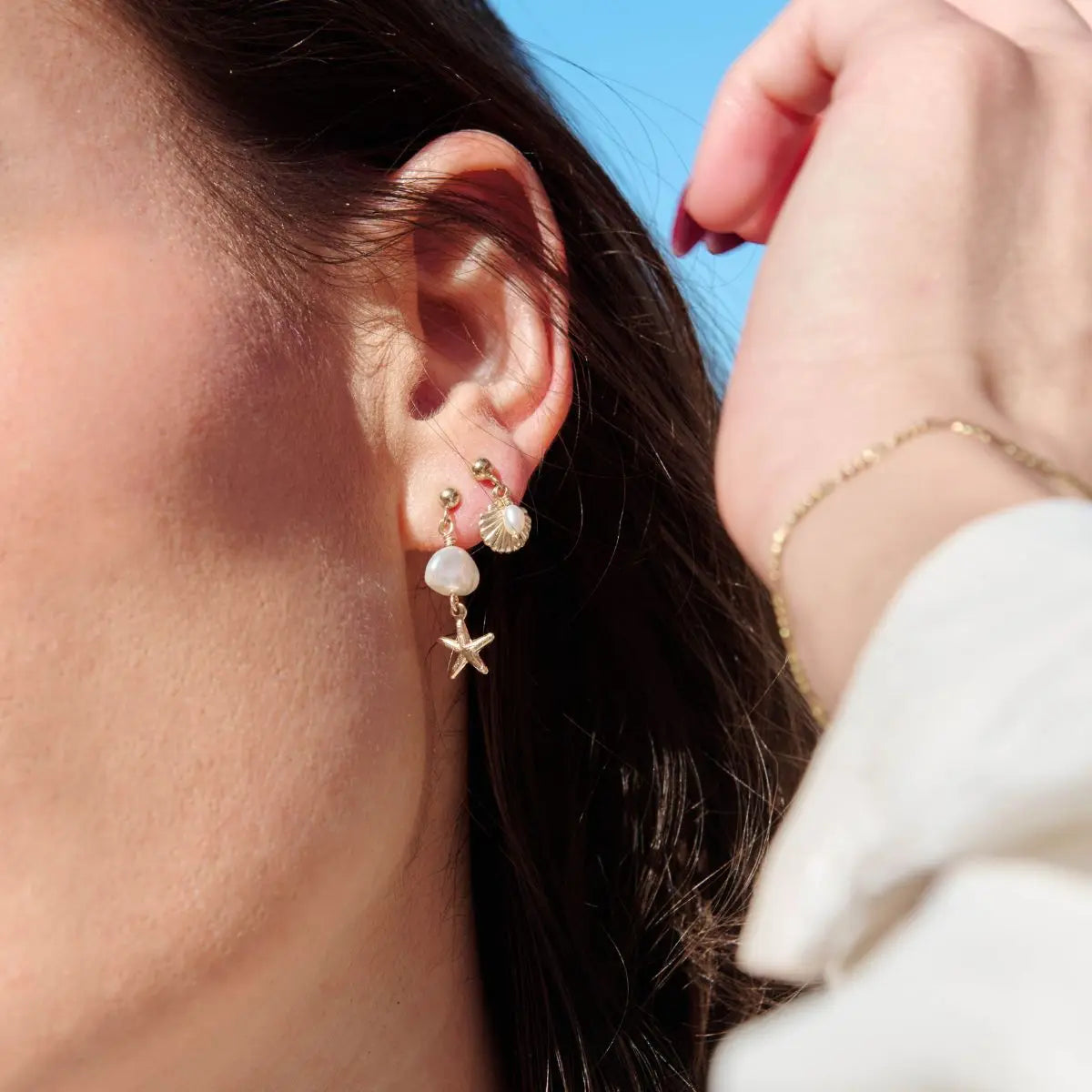 Close-up of an ear wearing a pearl and starfish earring with a hand touching the hair against a blue sky.
