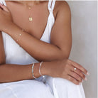 Close-up of a person wearing gold jewelry including a necklace, bracelet, and ring on a neutral background.
