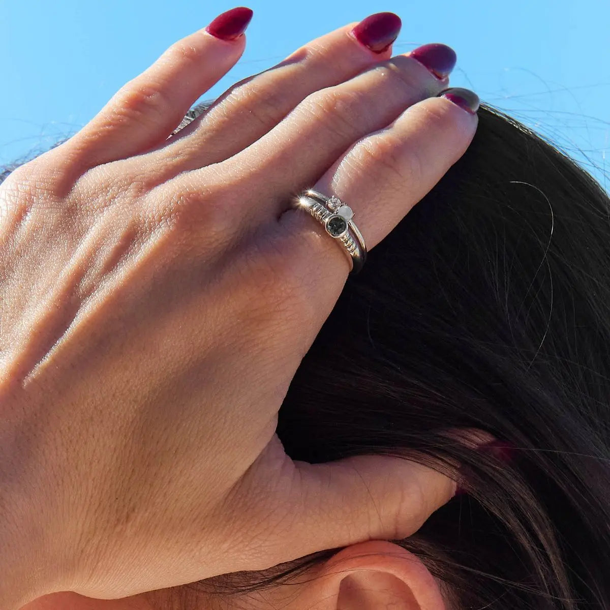 Close-up of a hand with red nail polish and a silver ring, holding hair against a blue sky.