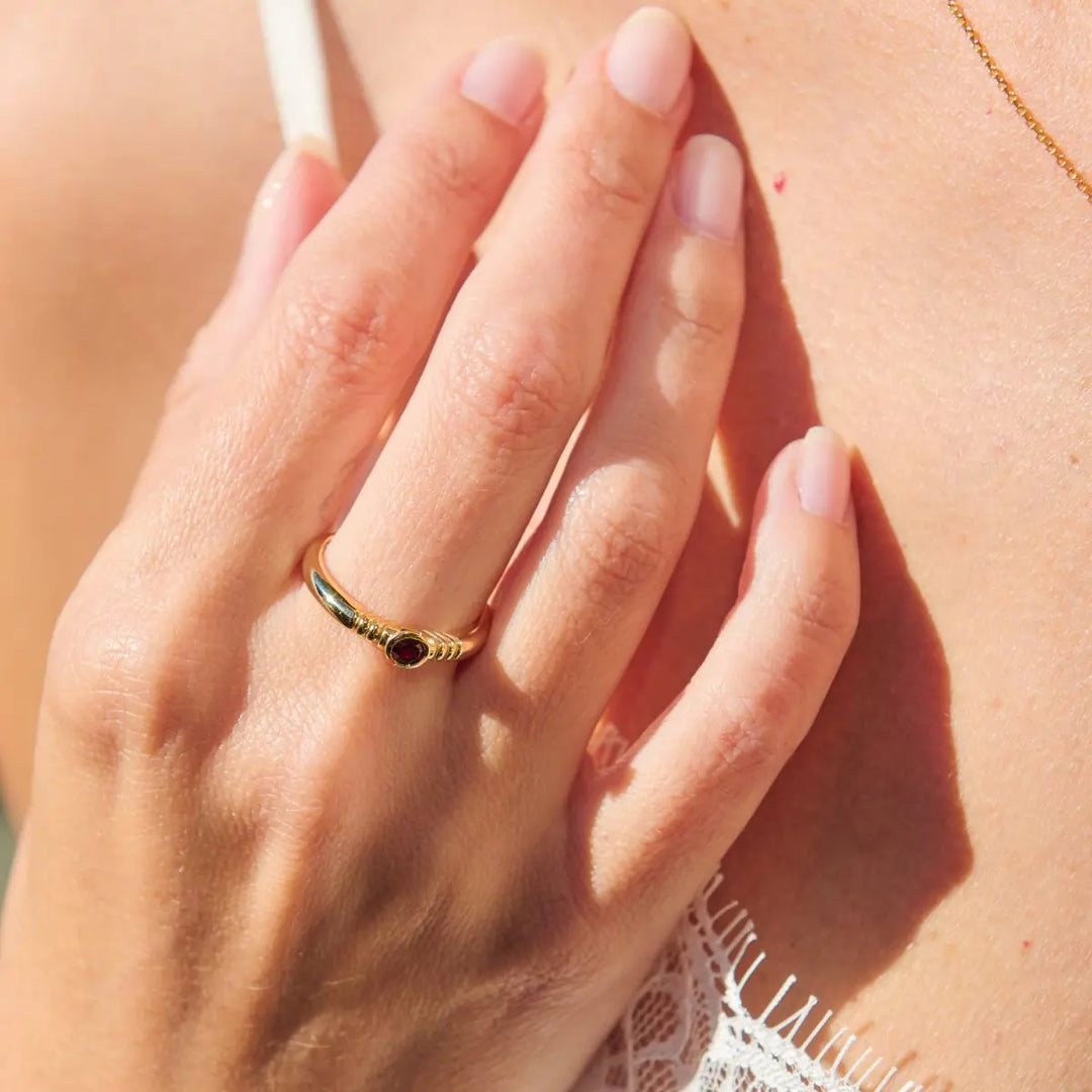 Close-up of a hand wearing a gold ring with a red gemstone, touching skin.
