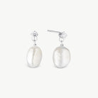 A pair of silver drop earrings featuring large, irregular white baroque pearls and a small clear crystal accent at the top, set against a plain off-white background.