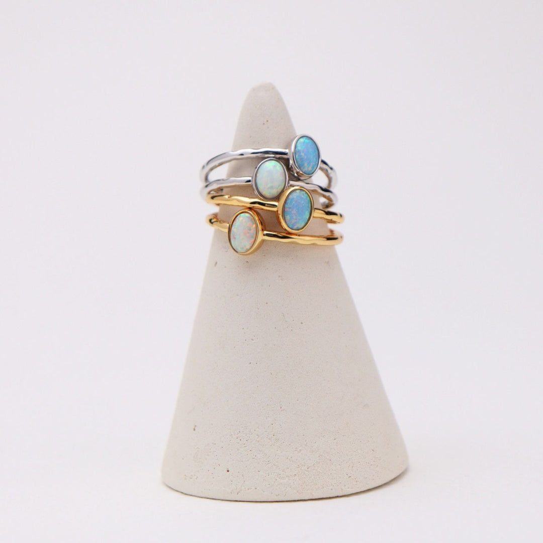 Three slim rings displayed on a cone-shaped jewelry stand against a clean white background. The rings feature oval cabochon stones in light blue and milky white, set in silver and gold-toned metal.