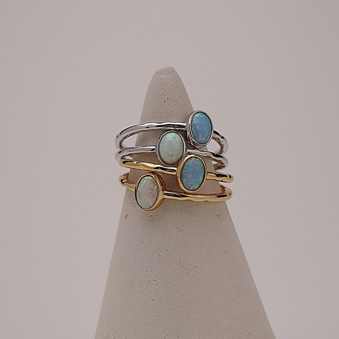 Three slim stacking rings displayed on a cone-shaped jewelry stand against a soft light gray background. Two rings are gold-toned with oval opal stones, and one ring is silver-toned with a larger oval opal stone.