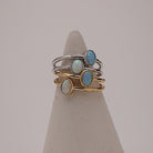 Three slim stacking rings displayed on a cone-shaped jewelry stand against a soft light gray background. Two rings are gold-toned with oval opal stones, and one ring is silver-toned with a larger oval opal stone.