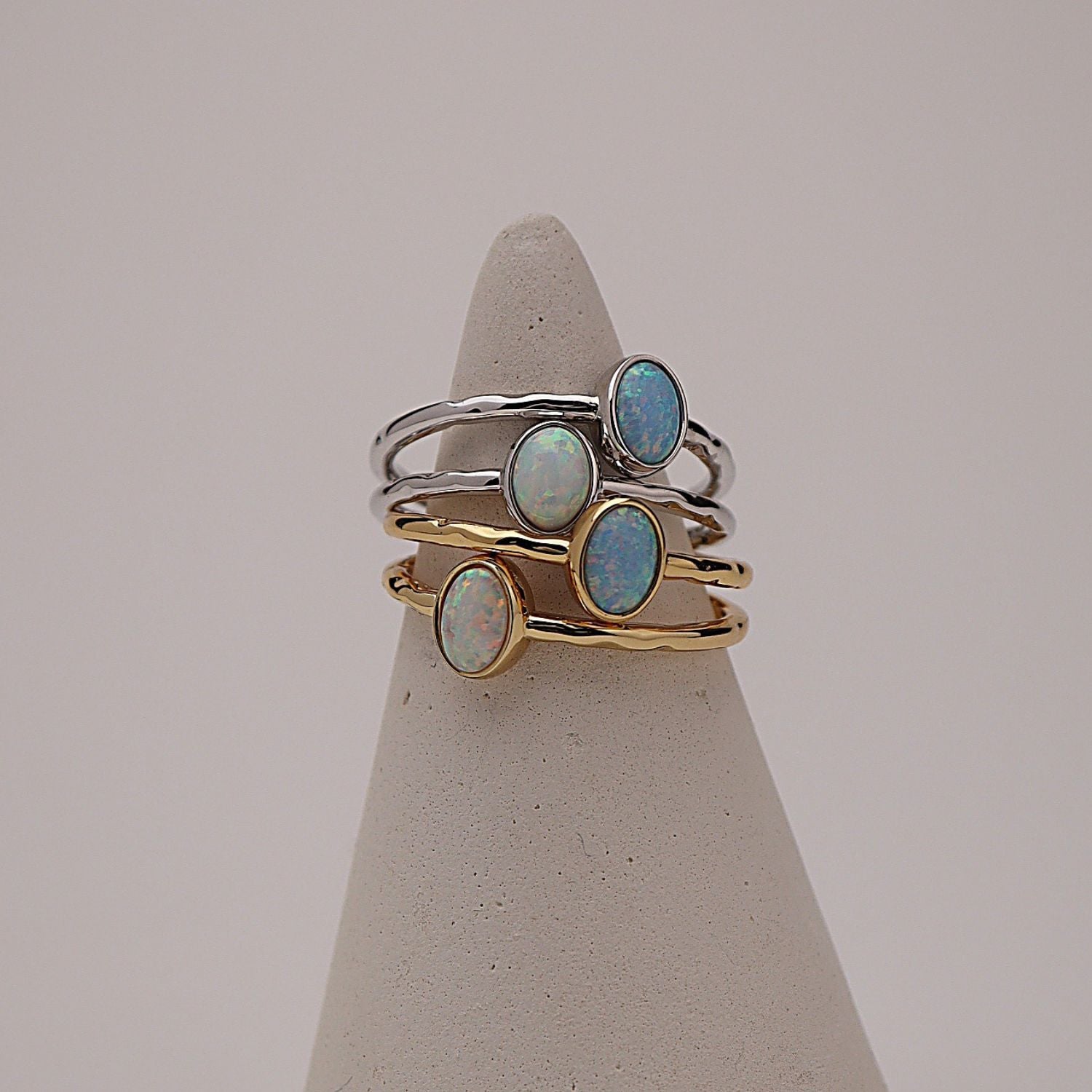 Three slim stacking rings displayed on a cone-shaped jewelry stand against a soft light gray background. Two rings are gold-toned with oval opal stones, and one ring is silver-toned with a larger oval opal stone.
