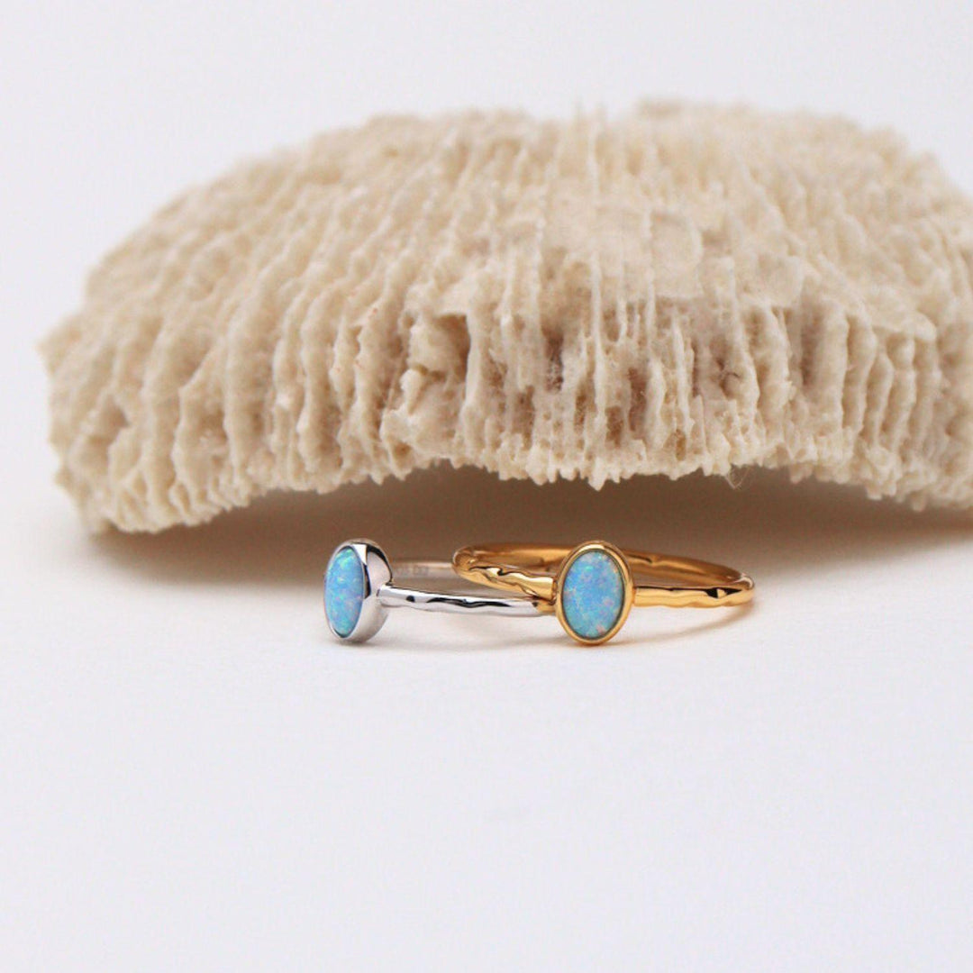 Two delicate rings with oval light blue opal stones, one in a yellow gold setting and the other in a silver-toned setting, displayed on a white surface with a textured beige coral prop in the background.