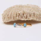 Two delicate rings with oval light blue opal stones, one in a yellow gold setting and the other in a silver-toned setting, displayed on a white surface with a textured beige coral prop in the background.