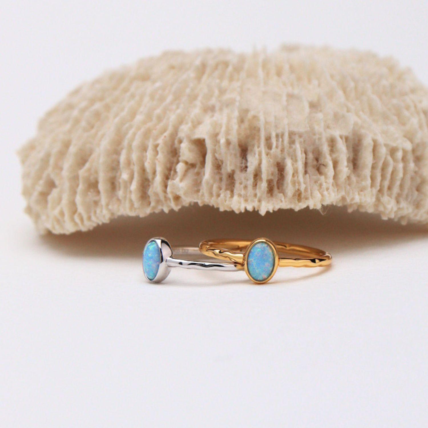 Two delicate rings with oval light blue opal stones, one in a yellow gold setting and the other in a silver-toned setting, displayed on a white surface with a textured beige coral prop in the background.
