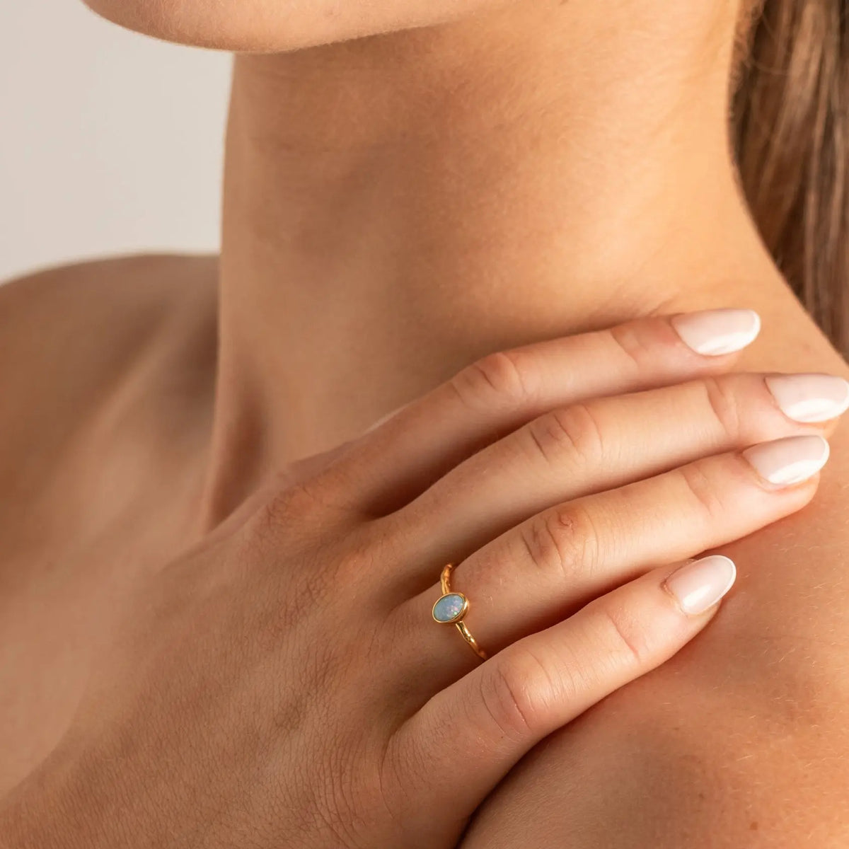 A close-up lifestyle shot of a woman's hand resting on her shoulder, wearing a delicate gold ring with a small oval light blue stone.
