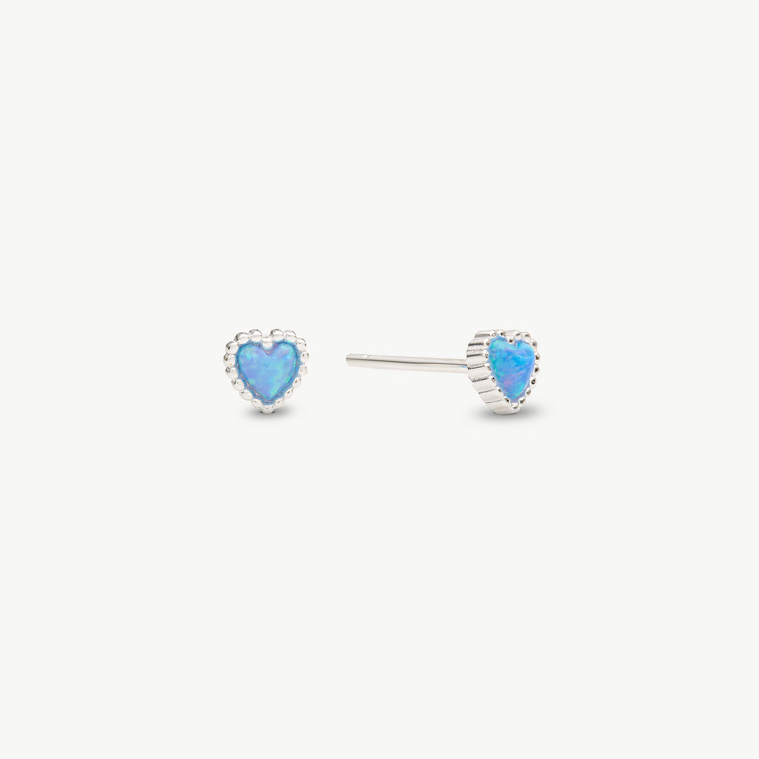 A pair of silver heart stud earrings with vibrant blue opal centers, shown on a clean off-white background.