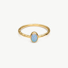 A gold ring with a textured band and an oval-shaped light blue opal center stone, shown on a clean off-white background.
