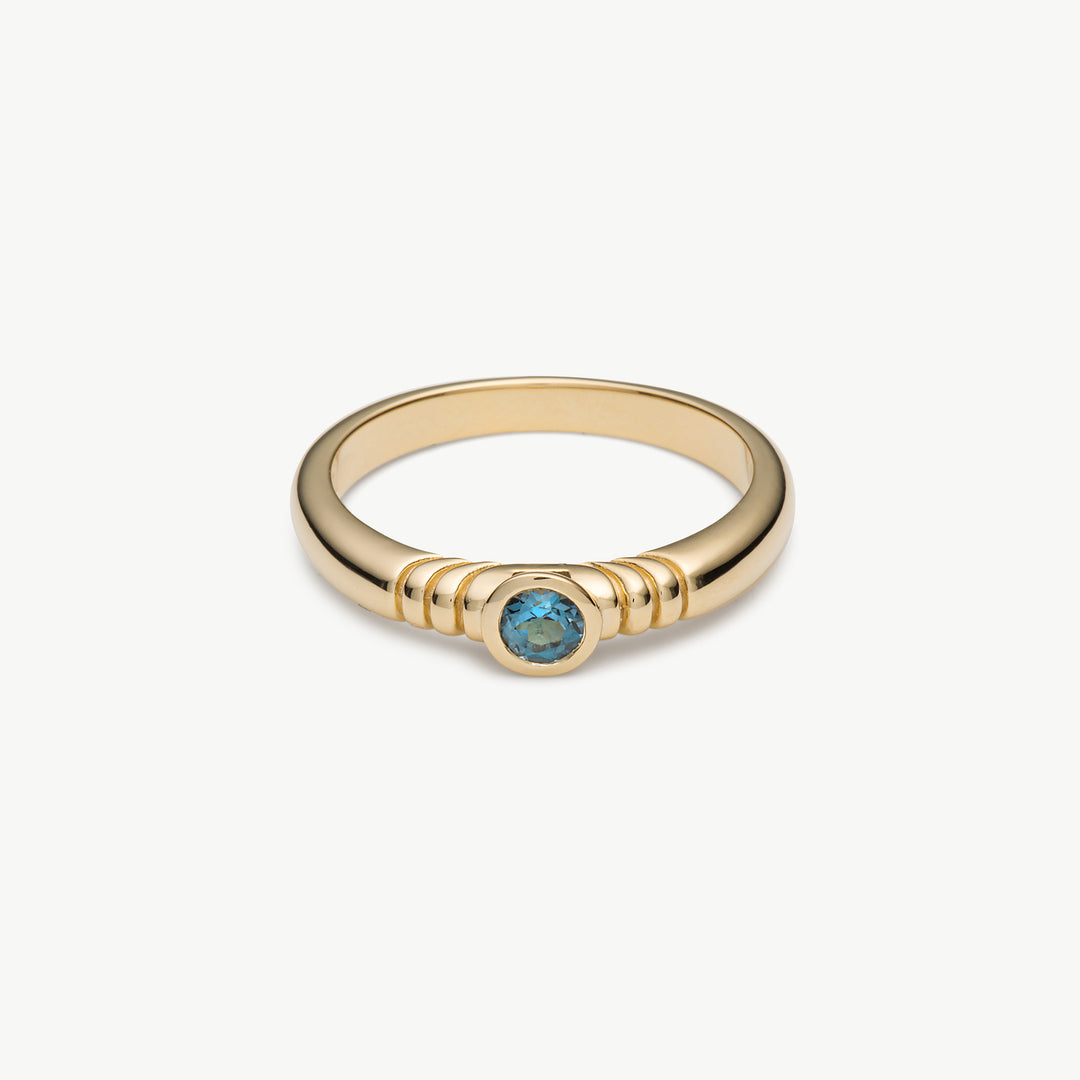 A gold ring with a textured band and a central round blue gemstone, shown on a clean off-white background.