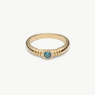 A gold ring with a textured band and a central round blue gemstone, shown on a clean off-white background.