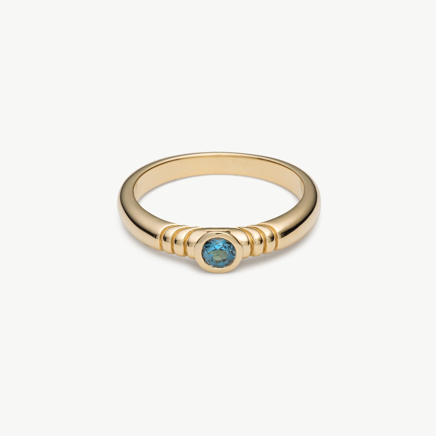 A gold ring with a textured band and a central round blue gemstone, shown on a clean off-white background.