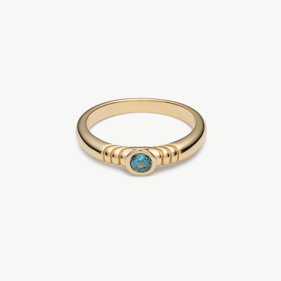 A gold ring with a textured band and a central round blue gemstone, shown on a clean off-white background.