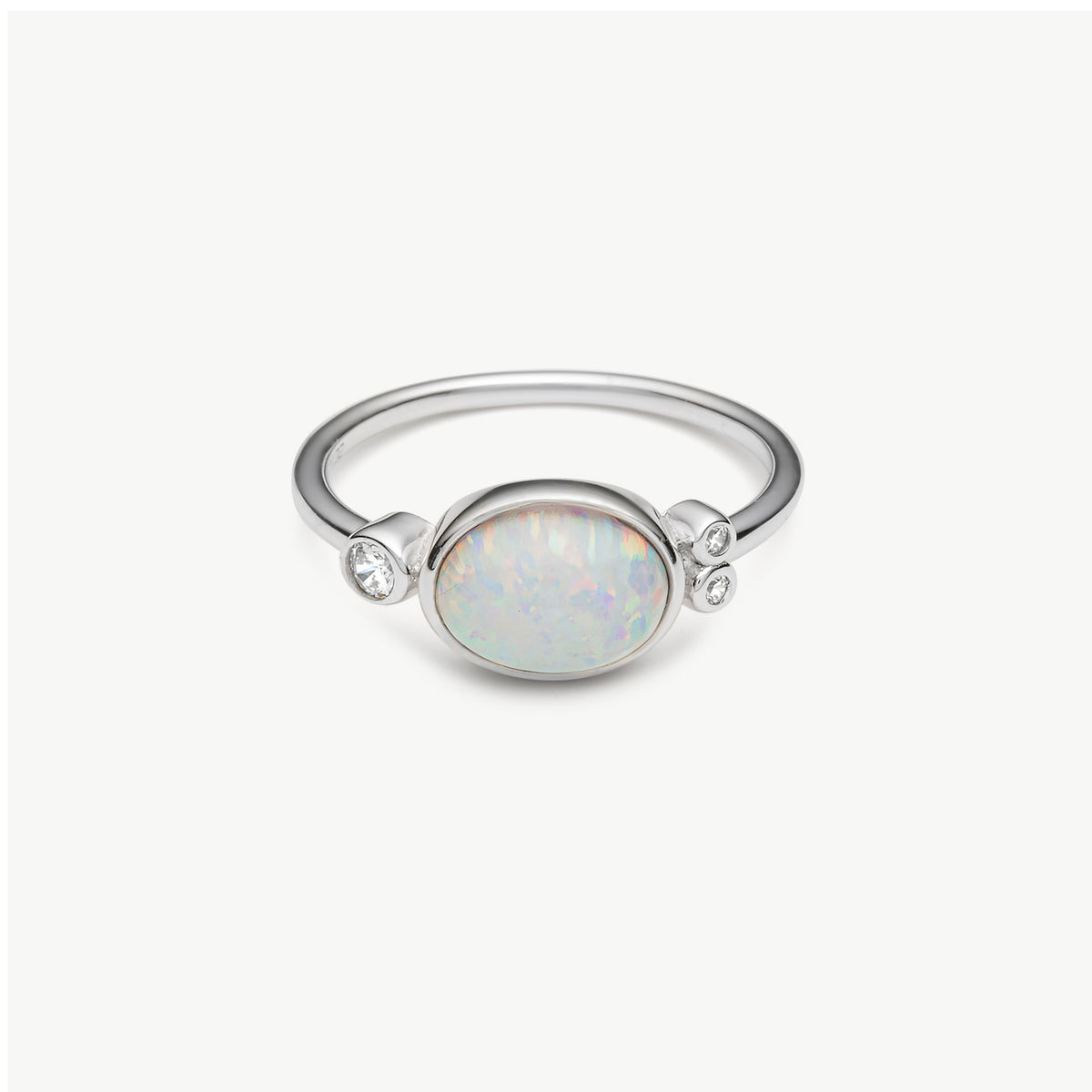 A silver ring featuring an oval cabochon opal with iridescent white and pastel colors, accented by two small clear stones on either side of the band, set against a clean off-white background.