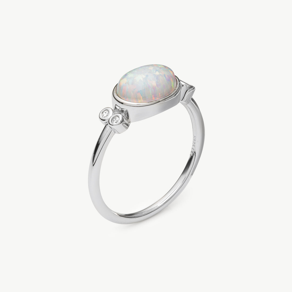 A silver ring featuring an oval cabochon opal with iridescent white and pastel colors, accented by two small clear stones on either side of the band, set against a clean off-white background.
