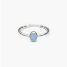 A silver ring with a textured band and an oval cabochon opal gemstone featuring blue, pink, and white play-of-color, set against a clean off-white background.