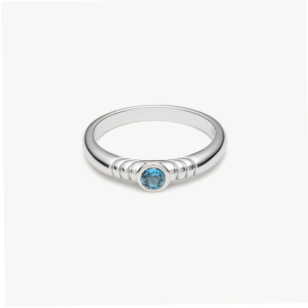 A silver ring with a textured band and a central round-cut light blue gemstone, shown on a clean off-white background.