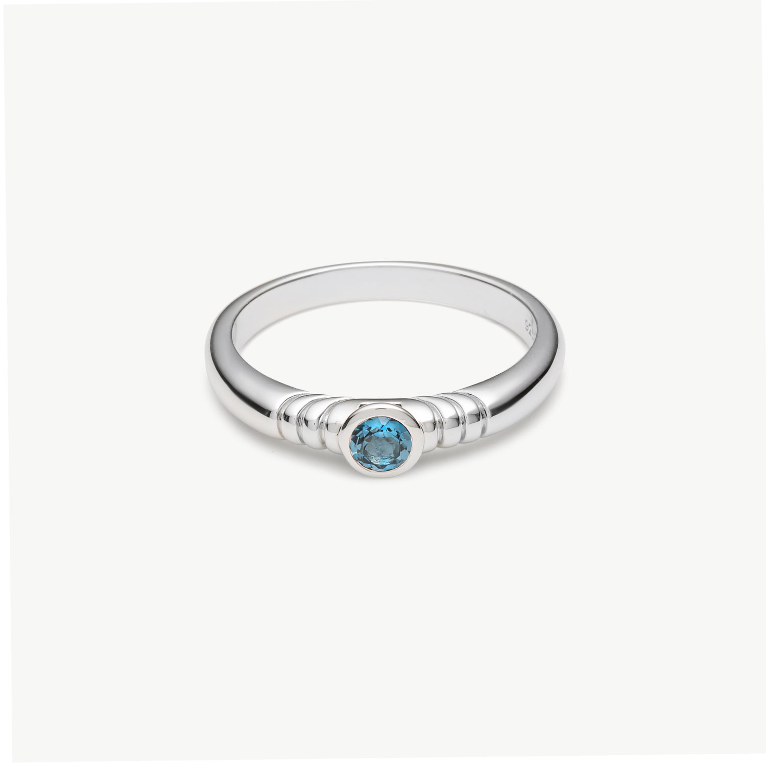 A silver ring with a textured band and a central round-cut light blue gemstone, shown on a clean off-white background.