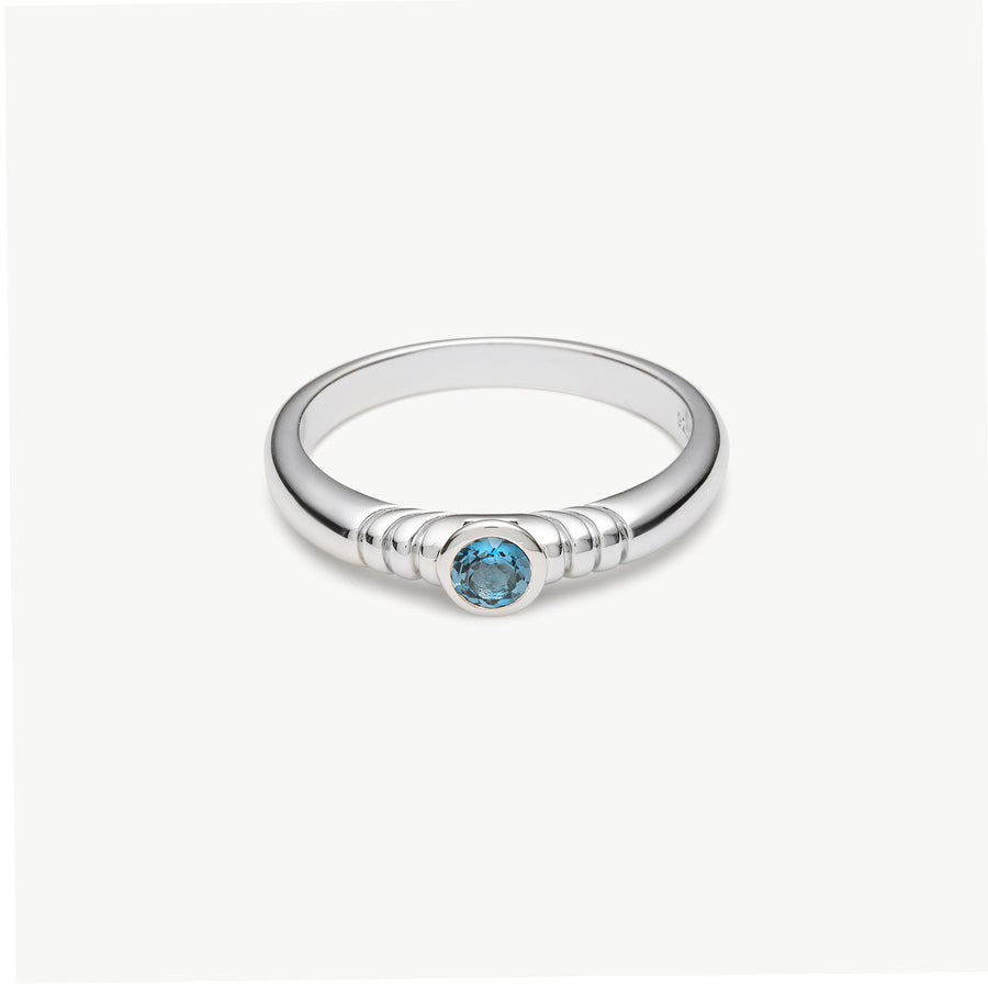 A silver ring with a textured band and a central round-cut light blue gemstone, shown on a clean off-white background.