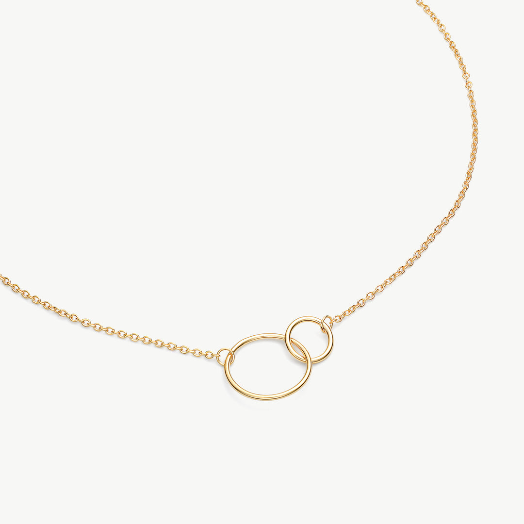A delicate gold chain necklace featuring two interlocking oval rings at the center, displayed on a clean off-white background.