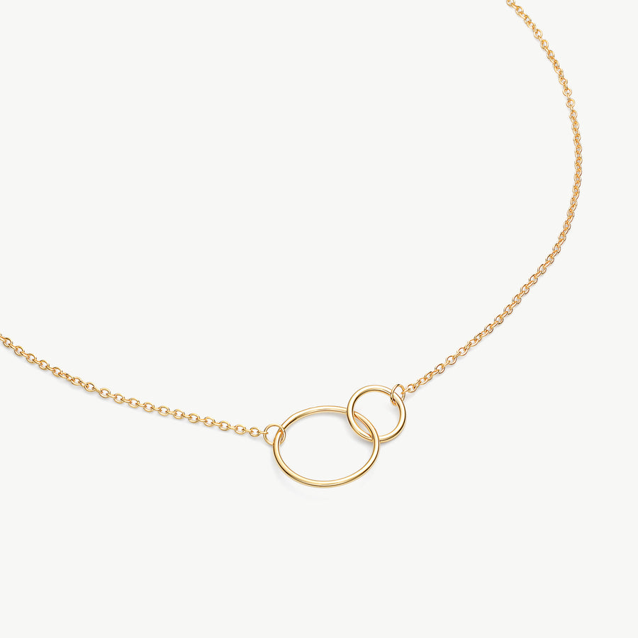 A delicate gold chain necklace featuring two interlocking oval rings at the center, displayed on a clean off-white background.