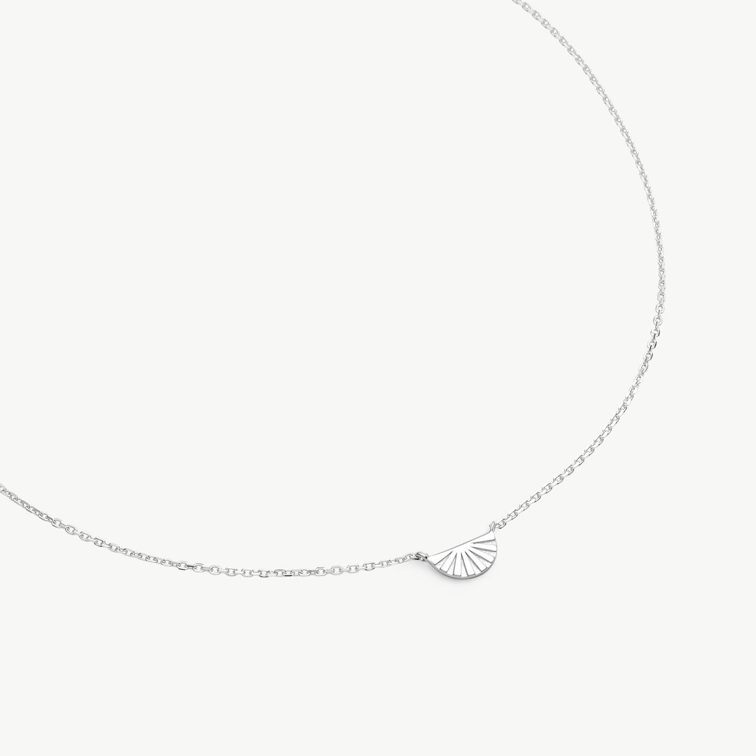A delicate silver chain necklace featuring a small, fan-shaped shell pendant, displayed against a clean, light off-white background.