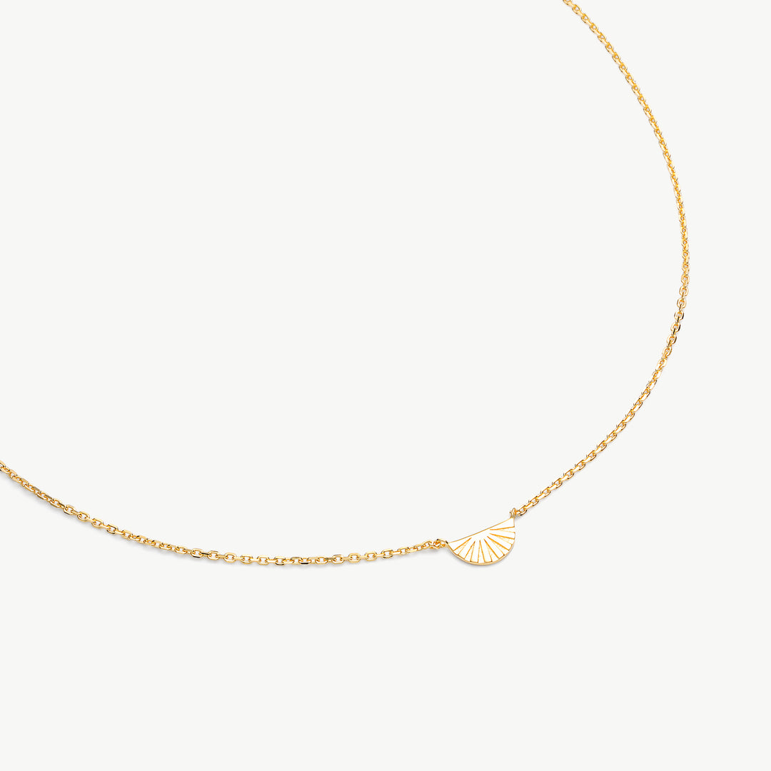 A delicate gold chain necklace featuring a small, minimalist fan-shaped pendant with radiating lines, displayed on a clean off-white background.