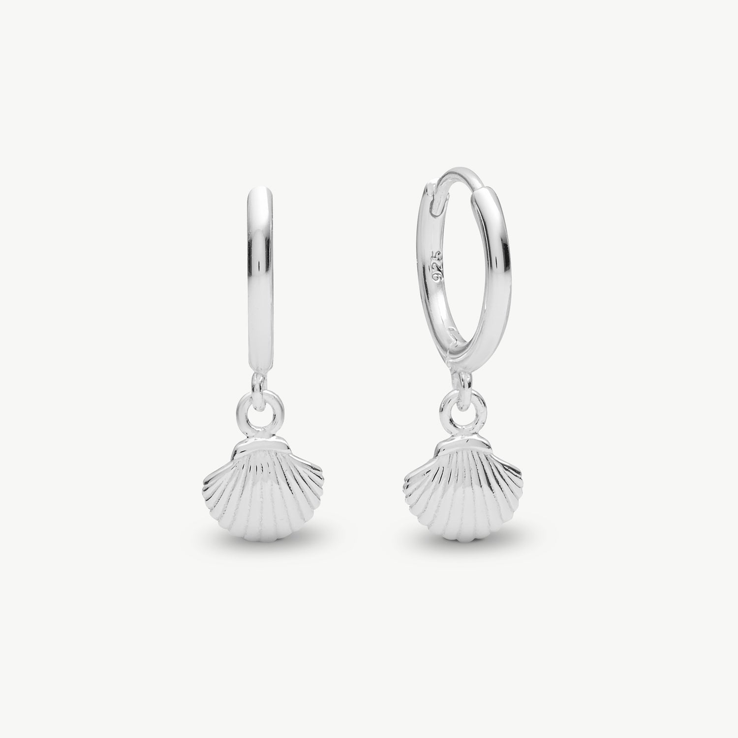 A pair of sterling silver huggie hoop earrings with dangling scallop shell charms, shown on a light gray background.